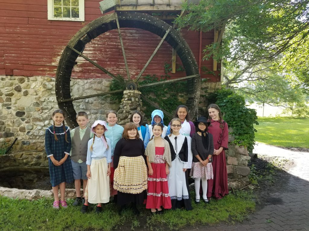 students dressed up in history outfits standing in front of a water wheel